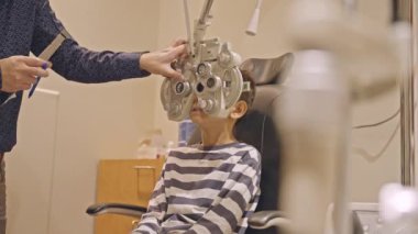 Küçük çocuklar göz muayenesi için göz doktoruna gittiklerinde phoropter kullanmışlar. Doktor elleri phoropter 'i genç çocuğun gözlerine yaklaştırıyor.