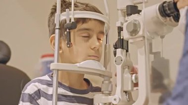 Küçük çocuklar göz muayenesi için göz doktoruna gittiklerinde phoropter kullanmışlar. Doktor elleri phoropter 'i genç çocuğun gözlerine yaklaştırıyor.