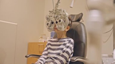 Küçük çocuklar göz muayenesi için göz doktoruna gittiklerinde phoropter kullanmışlar. Doktor elleri phoropter 'i genç çocuğun gözlerine yaklaştırıyor.