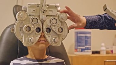 Küçük çocuklar göz muayenesi için göz doktoruna gittiklerinde phoropter kullanmışlar. Doktor elleri phoropter 'i genç çocuğun gözlerine yaklaştırıyor.