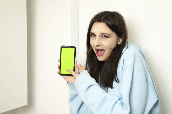 The model points to the green screen of the phone. Emotional girl holding a phone in her hands where is the place for the inscription