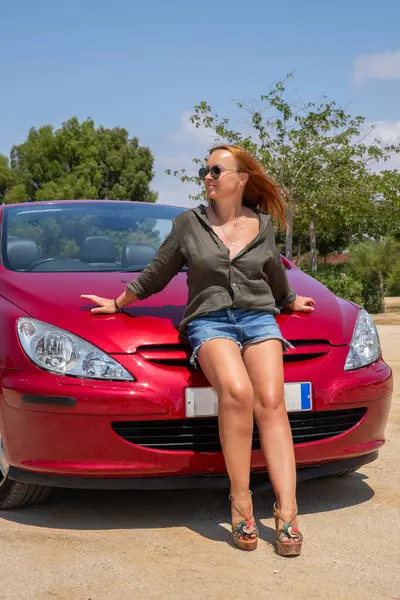 Kızıl saçlı kadın kırmızı arabayla tatilin tadını çıkarıyor.