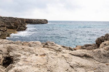 Bulutlu bir gökyüzünün altında, dalgalarla kaplı engebeli kayalık kıyı şeridi, okyanusun doğal güzelliğini vurguluyor ve düşünmek için huzurlu bir kıyı manzarası yaratıyor. Menorca 08.03.2025