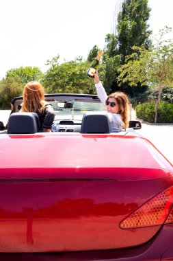 Kırmızı üstü açık bir arabada bir şişe şampanyayla neşeli kızlar.