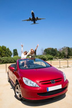 Arabadaki kız arkadaşlar uçan bir uçağın altında yaz tatiline hazırlanıyorlar. Kızlar üstü açık kırmızı bir arabada eğleniyor.
