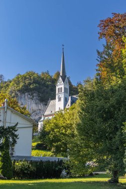 Lake Bled, Slovenia. St. Martin Church with gray-white mosaic roof surrounded by green foliage under blue cloudscape. White cliffs in back. Large white parish residential and office. clipart