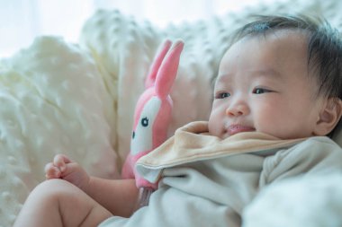 Pembe doldurulmuş bir oyuncakla oynarken yumuşak, rahat bir battaniyenin içinde rahatlatıcı bir görüntü. Bebeğin oyuncakla etkileşime girerken takındığı garip ifade çocukluk ve keşif döneminin güzel bir anını yakalar..