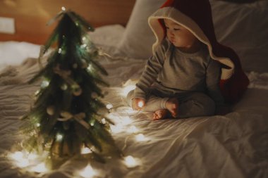 Kırmızı kapüşonlu battaniyeye sarılı tatlı bir bebek, sıcak bir yatakta oturuyor, yavaşça parlayan bir Noel ışığı tutuyor. Bebek meraklı bakışları, festival ışıklarının ve dekorasyonların sıcak, sihirli atmosferiyle çevrili, bayram mevsiminin harikasını yakalıyor.