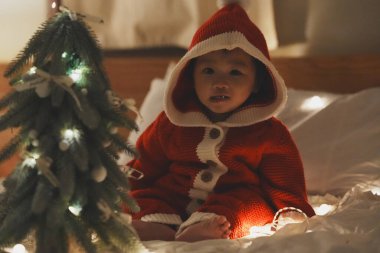 Kırmızı Noel Baba tarzı kapüşonlu kazak giymiş şirin bir bebek rahat bir yatakta oturur, ışıklarla süslenmiş küçük bir Noel ağacına bakar. Peri ışıklarının sıcacık parıltısı büyülü bir tatil ortamı yaratıyor, festival sezonunun neşesini ve mucizesini yakalıyor.