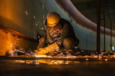Erkek işçi metali tank alt çelik plaka üzerinde kıvılcımlar kesiyor. Yakın çekim ışığı parlıyor. Koruyucu eldiven takıyor ve kapalı alanda maske takıyor..