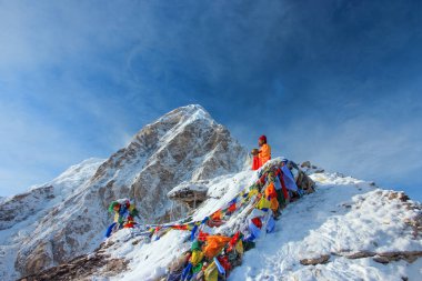 Himalayas, Nepal, May 10, 2016 - A monk with a bowl in his hands prays and meditates among colorful Buddhist prayer flags in the snow-capped mountains of Nepal.  clipart