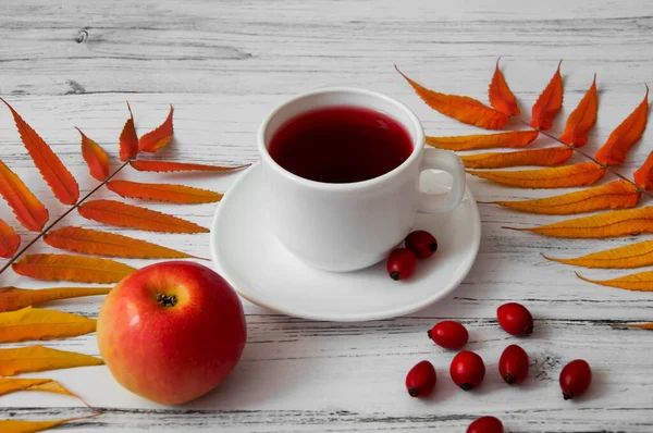 White cup of natural tea with rose hips on a wooden background with autumn leaves and apple. Autumn lifestyle