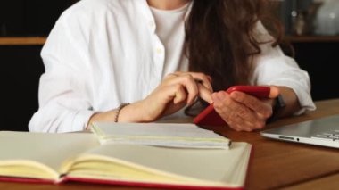 Cropped young brunette woman using mobile phone texting with friend, counting payments on digital calculator, sitting on table with laptop and notebooks at home office. Pay domestic bills fees online.