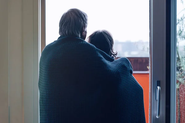 back view of a senior couple wrapped together in a blue blanket looking outside the terrace door in a rainy day