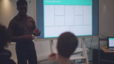 young professional of color stands in front of a group of colleagues, giving directions and explanations while using a TV monitor and laptop to display slides. He is specifically teaching the use of a business model canvas and strategies 