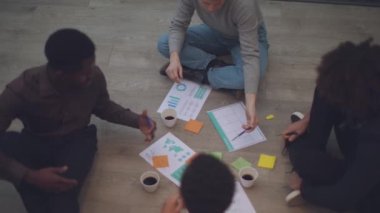 A top view of four young creatives and professionals sitting in a circle on the floor of an office. In the center, there are sheets of paper with charts, coffee in paper cups, colored pens, and post-it notes
