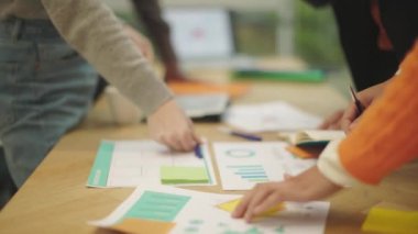 The table is cluttered with sheets of paper with charts, post-it notes, pens, coffee cups, notebooks, and a laptop. The shot captures the hands of different people with different skin tones, belonging to different ethnicities working together