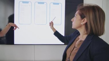Bir iş toplantısında iki profesyonel kadın, bir tanesi dijital ekrandaki kablo tasarımlarına işaret ediyor, üç farklı akıllı telefon uygulama düzeni sergiliyor - dijital tasarım incelemesi