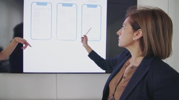 在商业会议上 两位专业女性 一位指向数字显示屏上的线框设计 展示了三种不同的智能手机应用程序布局 数字设计评审 — 图库视频影像