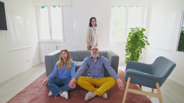 瞑想スタジオ 日本のセラピストは ソファーに対して床に座って瞑想する2人のコーカサス人の背後に立っています — ストック動画