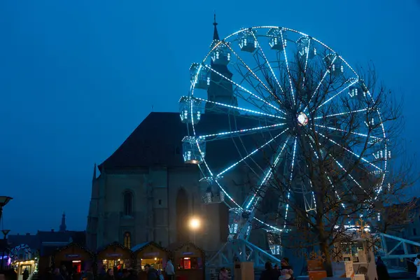 Cluj-Napoca, Romanya. 21 Aralık 2023: Cluj-Napoca Noel Pazarı 'nın dönme dolabı Romano-Katolik Kilisesi Aziz Michael' ın önünde, 1349-1480 yılları arasında, Cluj-Napoca 'nın merkezinde..