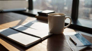 Pencerenin yanında dizüstü bilgisayarı, defteri ve kahvesi olan bir masa.