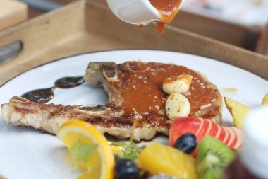 Lezzetli ızgara domuz pirzolası sarımsak, kahverengi sos ve yanında patates dilimleri, taze salata ve elma, kivi, portakal ve ejderha meyvesi gibi çeşitli meyvelerle servis edilir..