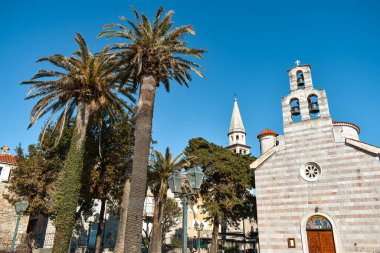Karadağ 'ın Budva kentindeki Aziz İvan ve Kutsal Üçleme Kilisesi. Katedralin yanındaki palmiye ağaçları, mavi gökyüzü, güneşli gün.