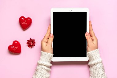 female hands holding digital tablet for chooses gifts, makes purchase, two red hearts on pink table Top view Flat lay Holiday shopping list, Happy Valentines day, party, online shop concept Mockup.