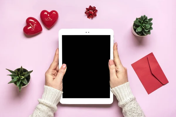 female hands holding digital tablet for chooses gifts, makes purchase, two red hearts on pink table Top view Flat lay Holiday shopping list, Happy Valentines day, party, online shop concept Mockup.