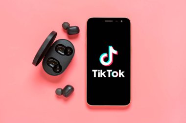 Bangkok, Thailand September 2022 Smartphone Tik Tok application icon, logo on screen, wireless headphones, charger capsule on pink table Trendy social media network concept Flat lay Top view.