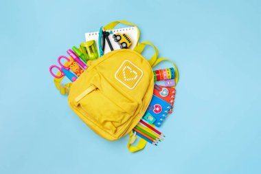 Okula dönmek, eğitim kavramı. Okul malzemeleriyle dolu sarı bir sırt çantası. Not defteri, kalemler, cetvel, hesap makinesi, makas mavi arka planda izole edilmiş. Üst Manzara. Düz dizilim bileşimini kopyala.