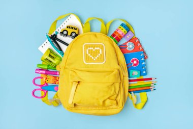 Okula dönmek, eğitim kavramı. Okul malzemeleriyle dolu sarı bir sırt çantası. Not defteri, kalemler, cetvel, hesap makinesi, makas mavi arka planda izole edilmiş. Üst Manzara. Düz dizilim bileşimini kopyala.