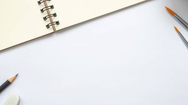 Beyaz arkaplanda boya fırçası, kalem ve silgi ile boş sarmal defteri aç, metnin için boşluk, çalışma alanının üst görünümü.
