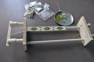 Özel bir dokuma tezgahında boncuklu bir bilezik hazırlayan yakın plan bir el. Bilezik renkli boncuklarla geleneksel bir desen içeriyor. Araçlar ve malzemeler düzenli olarak düzenlenir, titizlikle sergilenir, Skil.
