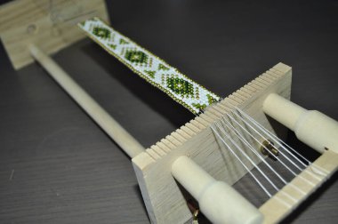 Özel bir dokuma tezgahında boncuklu bir bilezik hazırlayan yakın plan bir el. Bilezik renkli boncuklarla geleneksel bir desen içeriyor. Araçlar ve malzemeler düzenli olarak düzenlenir, titizlikle sergilenir, Skil.