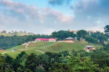 Bulutlu gökyüzünün altındaki yeşil bir dağda kırmızı renkli tatil köyü