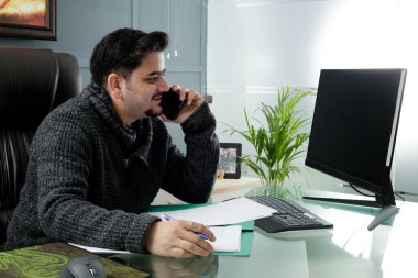 A young businessman is sitting in the office doing his work and talking on mobile phone.