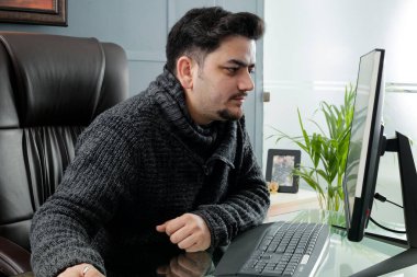A young businessman is sitting in the office working on computer and smiling.