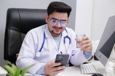 Genç bir doktor klinikte oturmuş su içiyor ve cep telefonundan bir şeyler izliyor..