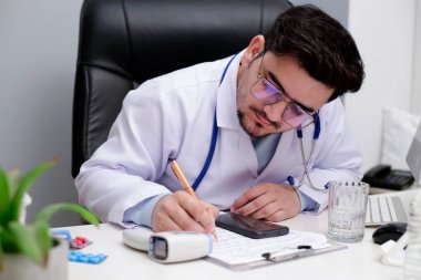 Genç bir doktor klinikte oturuyor ve kağıda kalemle bir şeyler yazıyor..