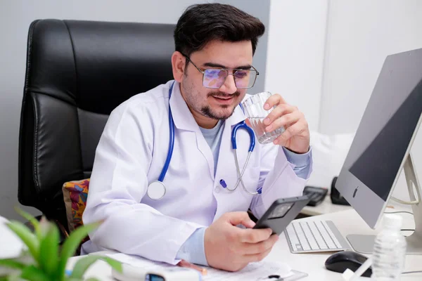 Młody Lekarz Siedzi Klinice Pije Wodę Ogląda Coś Komórce — Zdjęcie stockowe