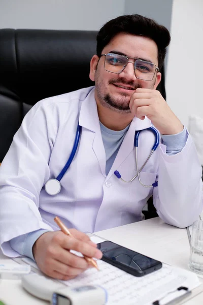 Młody Lekarz Siedzi Klinice Trzymając Długopis Dłoni Uśmiechając Się — Zdjęcie stockowe