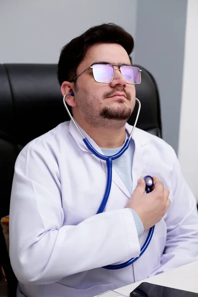 一位年轻的医生正坐在诊所里用听诊器检查他的心跳 — 图库照片