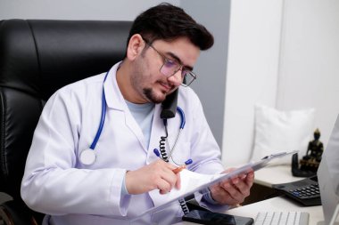 Genç bir doktor klinikte oturmuş telefonla konuşuyor ve rapor okuyor..