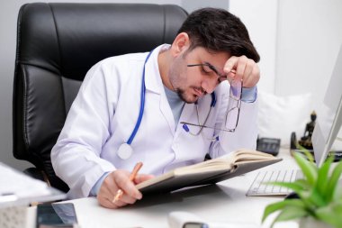 Genç bir doktor klinikte oturmuş günlükten bir şeyler okuyor ve yorgun hissediyor..