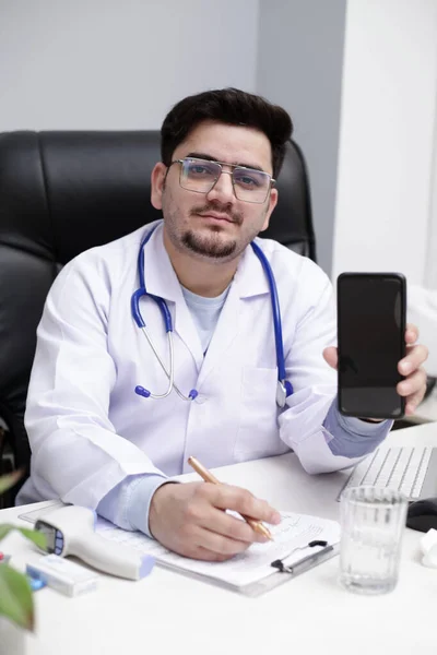 Młody Lekarz Siedzi Klinice Trzymając Ręce Telefon Komórkowy Pokazuje Coś — Zdjęcie stockowe