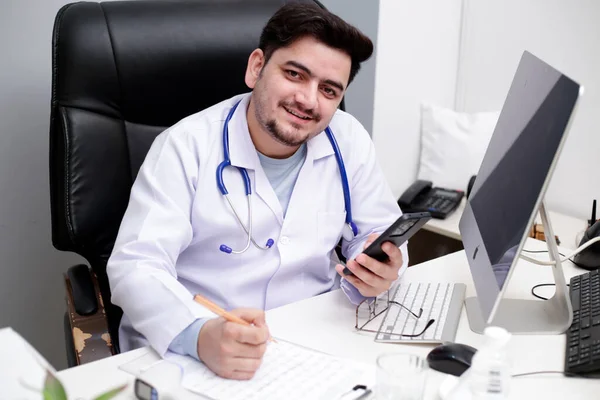 Młody Lekarz Siedzi Klinice Trzymając Telefon Ręce Uśmiechając Się — Zdjęcie stockowe