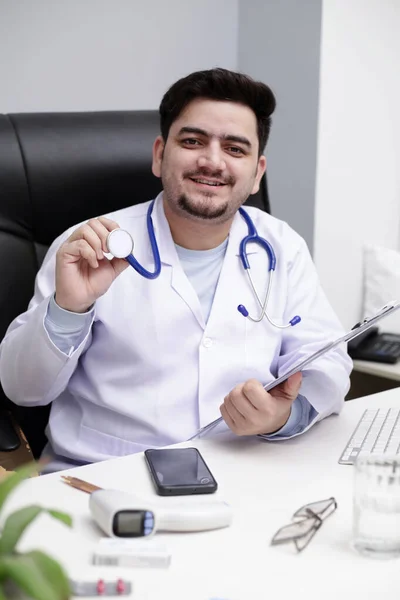 Młody Lekarz Siedzi Klinice Trzymając Stetoskop Dłoni Uśmiechając Się — Zdjęcie stockowe