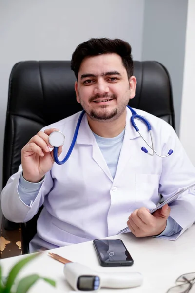Joven Médico Está Sentado Clínica Sosteniendo Estetoscopio Mano Sonriendo — Foto de Stock